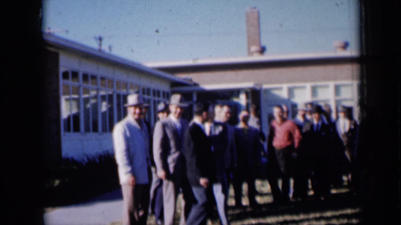 Several men walk past the camera while others stand near the building.