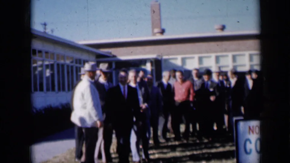 A man in a dark suit stands before a group of men outside the 1961 Missouri community building scene.