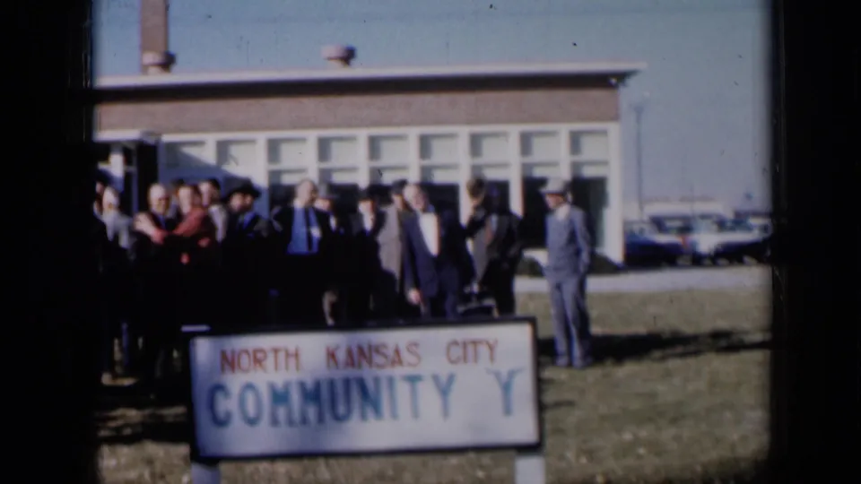 The group remains gathered outside the North Kansas City Community Y, with lawn, windows, and vintage cars visible in the 8mm frame.