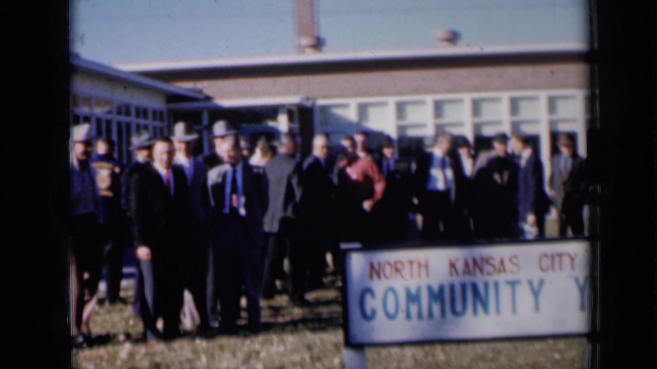 Men in suits and hats stand outside a building near a North Kansas City Community sign.