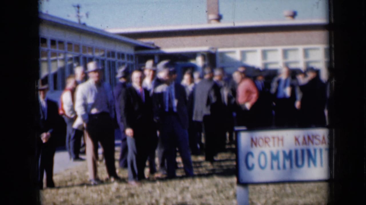 1961 Missouri footage frame with suited men and hats gathered on a lawn and sidewalk outside a low community building.