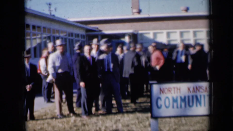 1961 Missouri footage frame with suited men and hats gathered on a lawn and sidewalk outside a low community building.