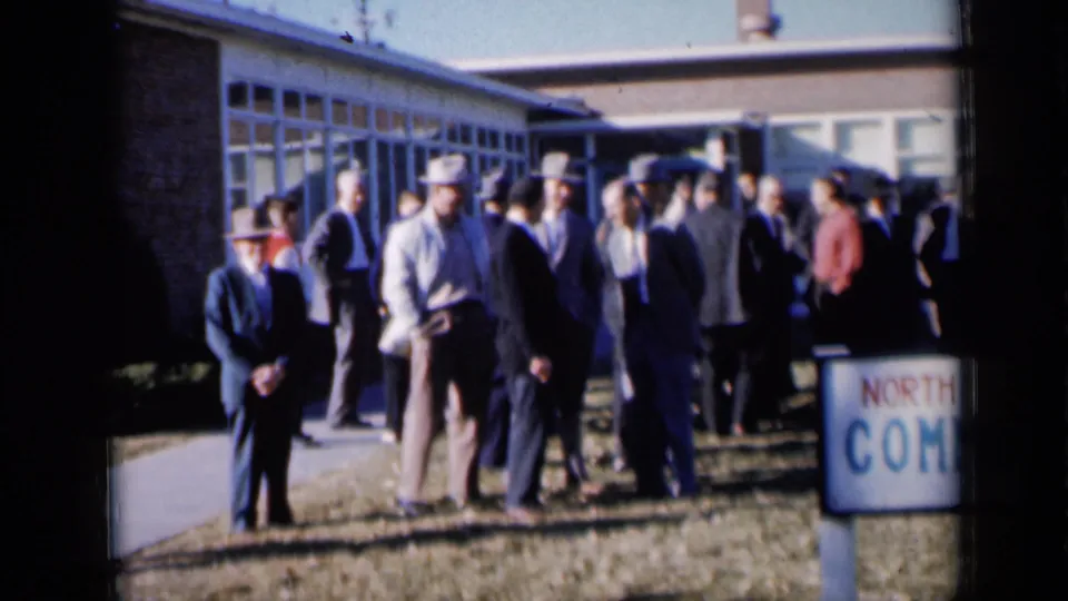 1961 Missouri home-movie frame of formally dressed men standing near the sidewalk outside a community building, with a partial North Kansas City sign.