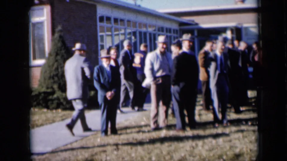 1961 Missouri frame showing men in suits, hats, and coats gathered casually on a lawn and walkway outside a community building.