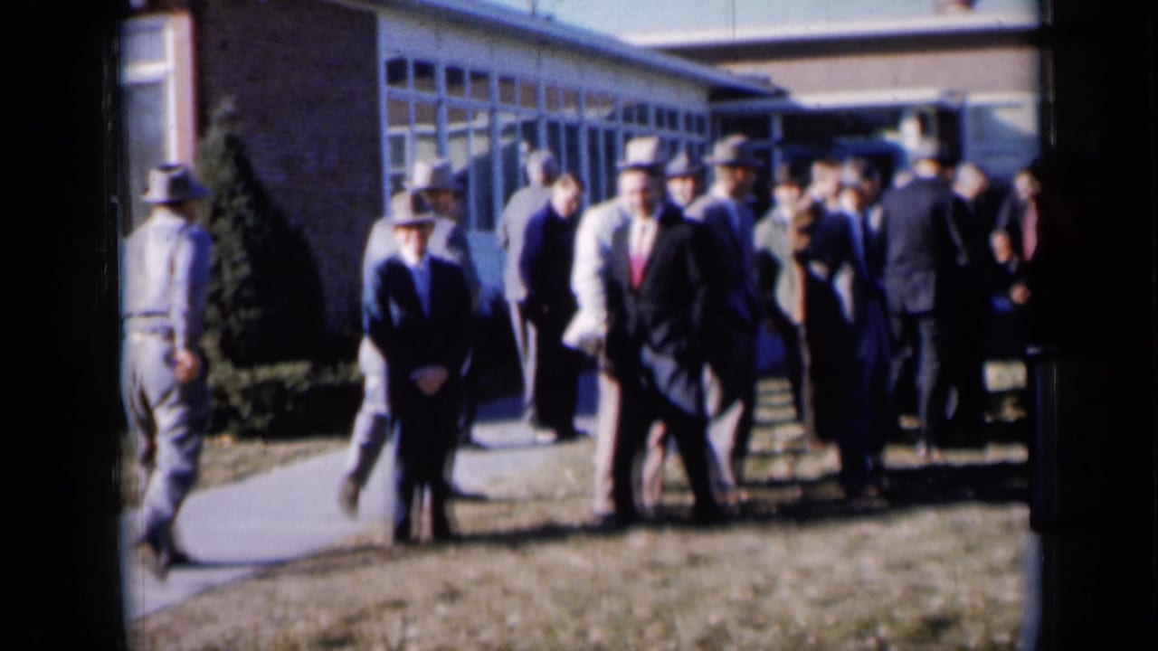 Men in suits and hats gather outside a low building on a sunny day.