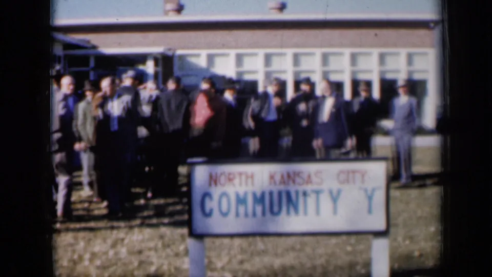 A crowd of suited men stands outside the North Kansas City Community Y, with the sign prominent in the foreground.