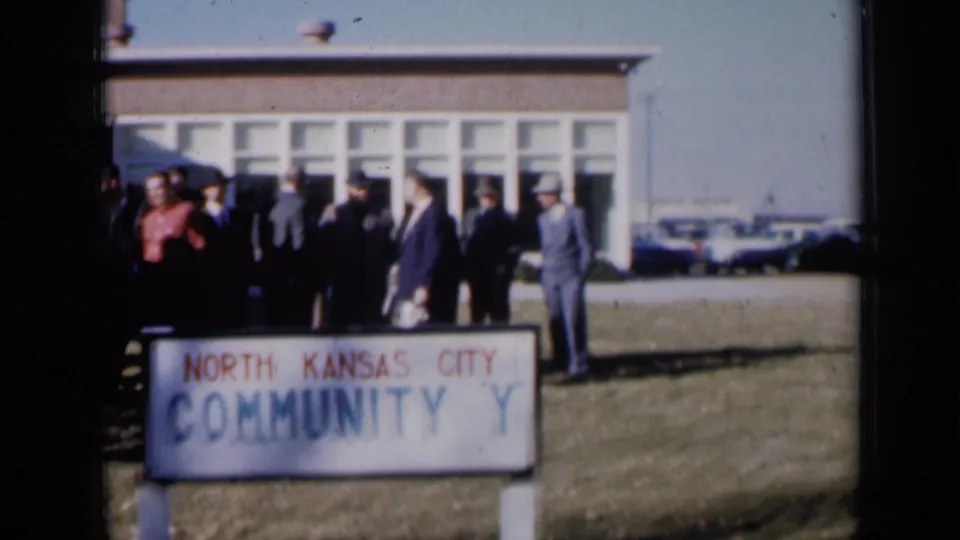 Wide 1961 Missouri frame with men near the North Kansas City Community Y sign, building windows, and parked cars.