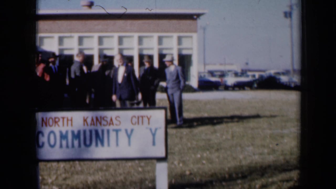 Men gather near a North Kansas City community sign outside a low public-looking building.