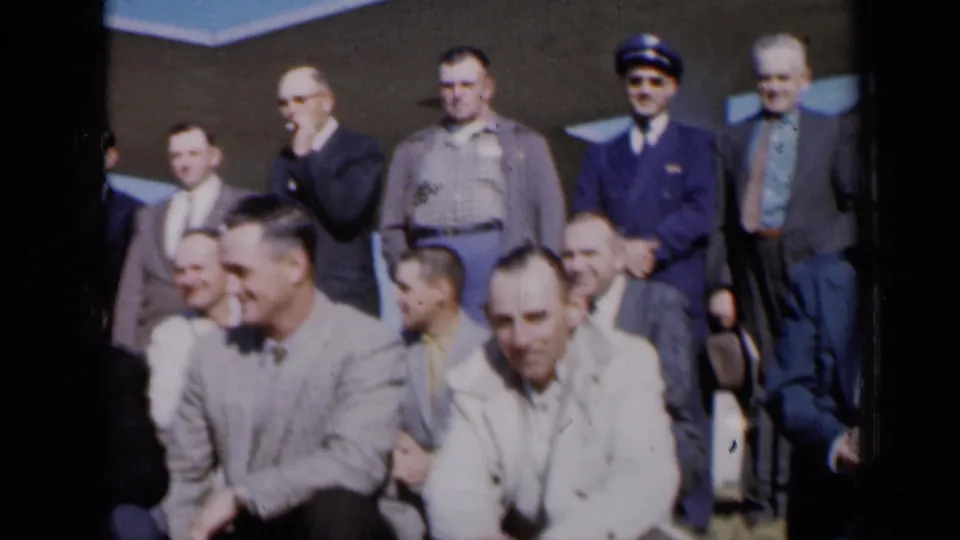 1961 Missouri frame of men in suits and jackets settling into a posed group outside a modern building.