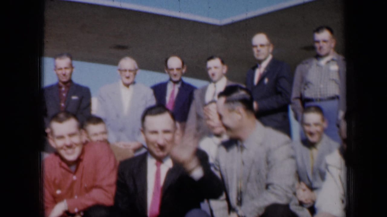 Adult men in suits and shirts pose outdoors beneath a concrete overhang.