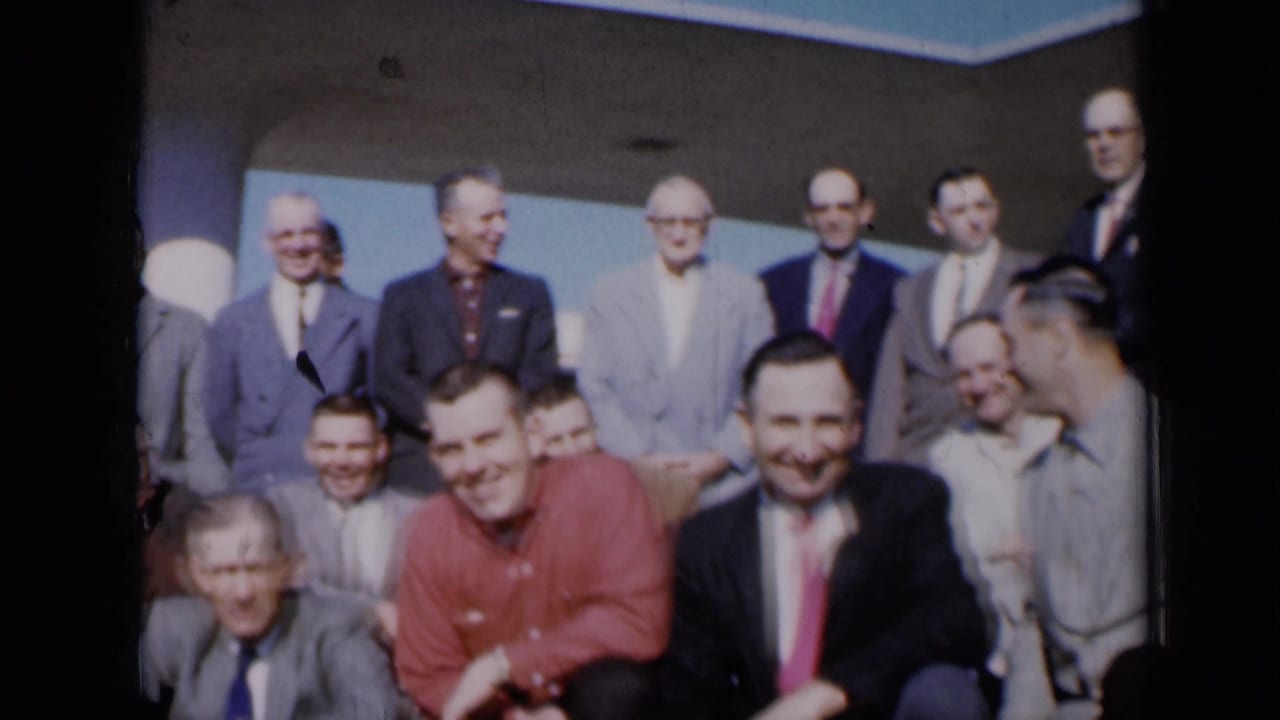 Group of adult men smile and pose in rows outside a building.