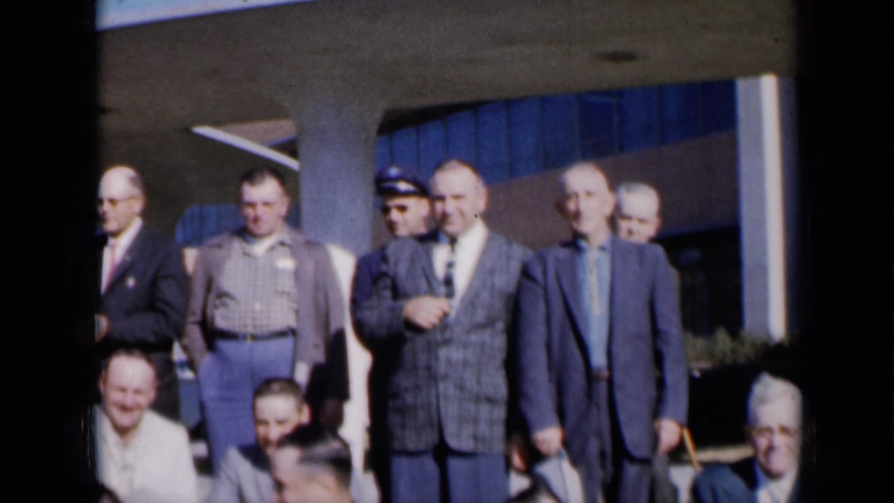 Men gathered outside a modern building, with some seated low in the foreground.