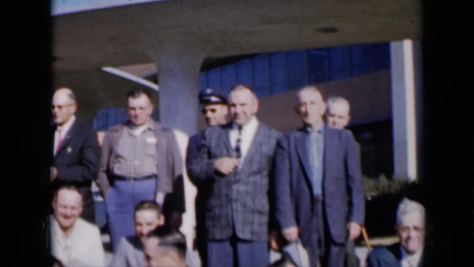 1961 Missouri home-movie frame showing adult men in suits and casual clothes outside a building with columns and blue windows.