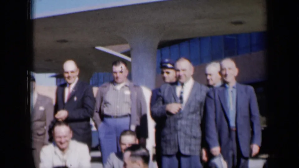 Adult men in 1960s clothing stand outside a Missouri building, with a plaid sport coat and uniform-style cap visible.