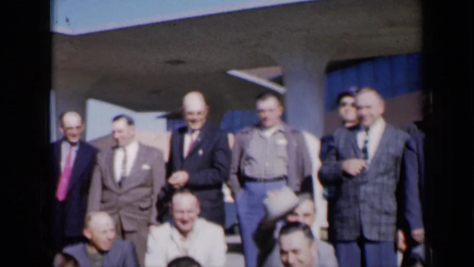 Candid 1961 Missouri frame of men laughing outside, with a hat being raised during a playful group moment.