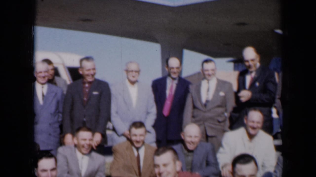 Men in suits gather in rows for a group photograph beside a white vehicle.