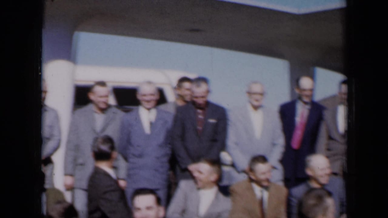 A group of men in suits stand and sit near a white vehicle and building columns.