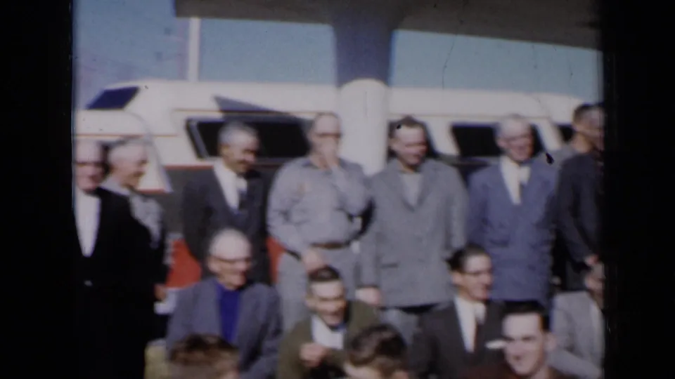 Mid-century dressed men, some wearing glasses, remain grouped in front of a white vehicle during the Missouri 1961 scene.
