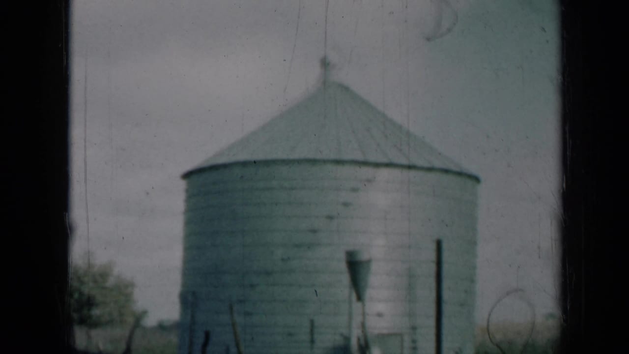 Large grey metal silo seen through dark vertical framing with pale sky behind it.