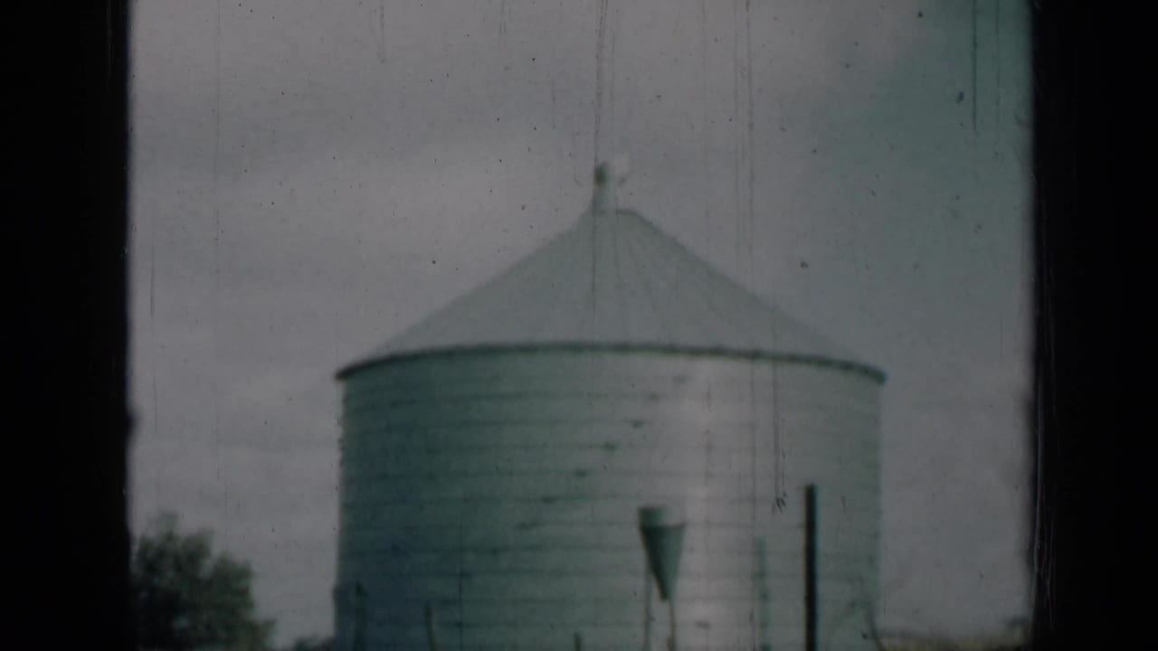 1961 Missouri frame of a large corrugated silo under a pale sky, with dark foreground framing and faint trees at the edge.