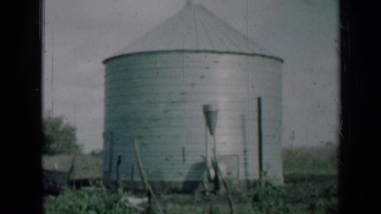 Final 1961 Missouri frame of the grey metal silo, foreground posts and low vegetation, with visible 8mm scratches and soft focus.
