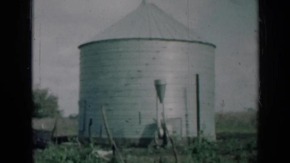 Final 1961 Missouri frame of the grey metal silo, foreground posts and low vegetation, with visible 8mm scratches and soft focus.
