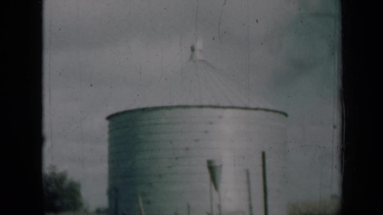 1961 Missouri farm view with a round metal silo, conical roof, foreground posts, grass, and muted home-movie color.