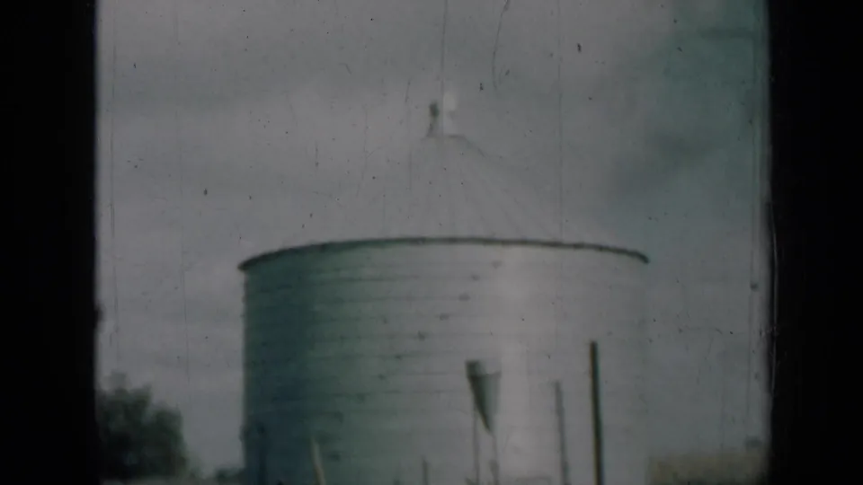 1961 Missouri farm view with a round metal silo, conical roof, foreground posts, grass, and muted home-movie color.