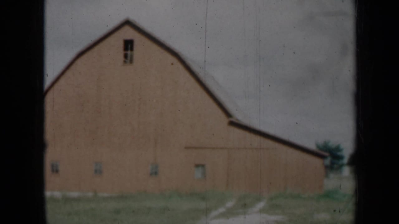 1961 Missouri home-movie frame of a barn exterior framed by dark window bars, with a pale dirt track, grass, trees, and cloudy sky.