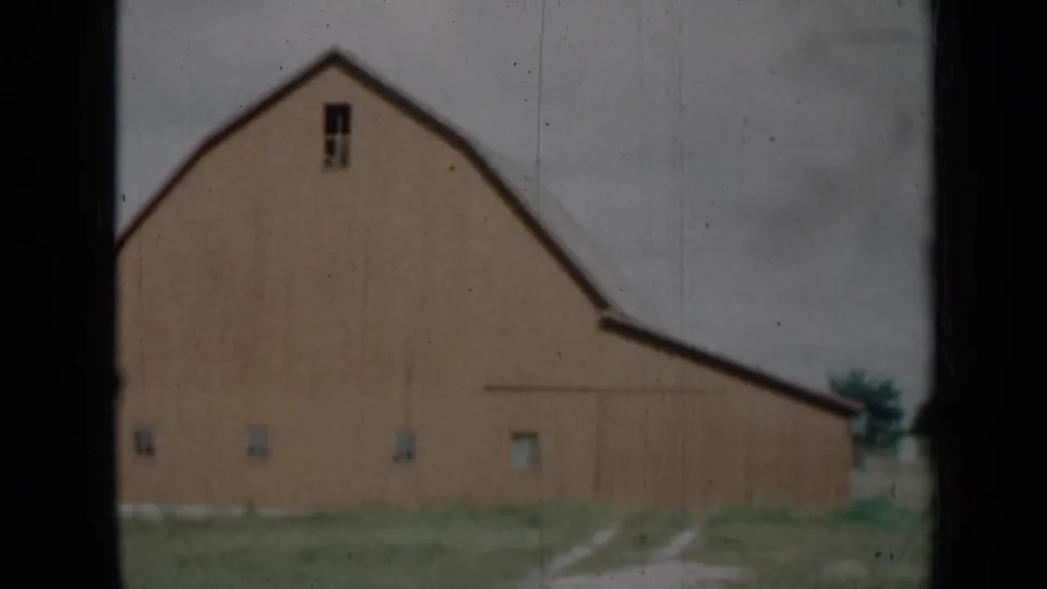 1961 Missouri home-movie frame of a barn exterior framed by dark window bars, with a pale dirt track, grass, trees, and cloudy sky.