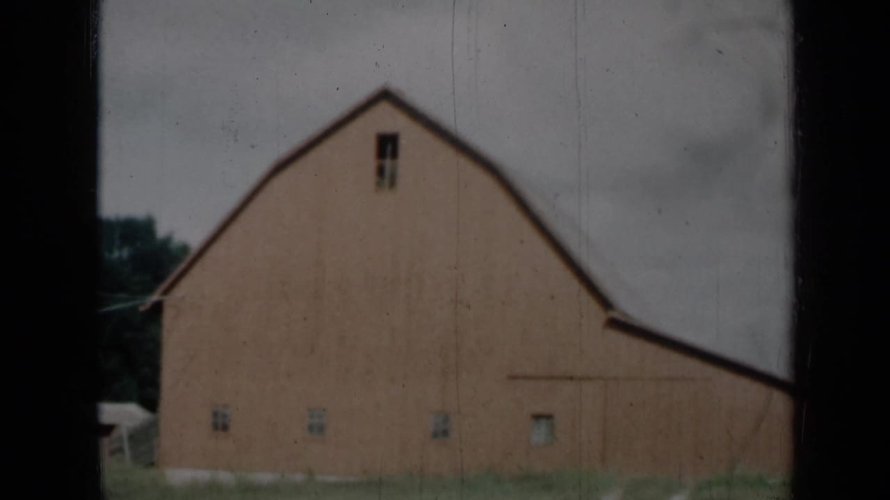 1961 Missouri frame showing the barn facade from a slightly shifted angle, with small windows, grass, trees, cloudy sky, and dark side framing.