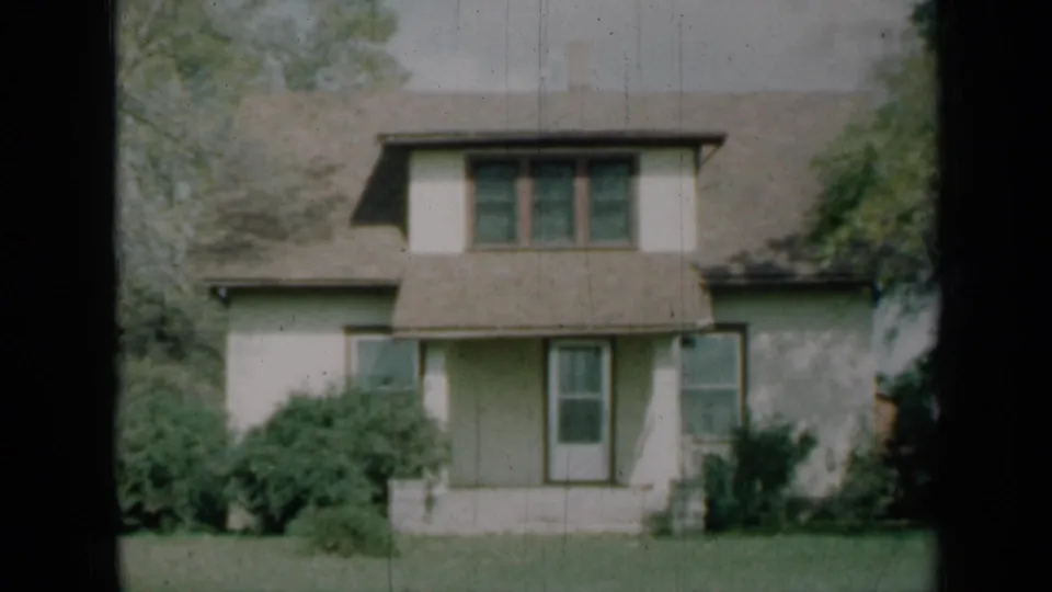 1961 Missouri home-movie view of a modest white house with brown trim, covered porch, shrubs and lawn.