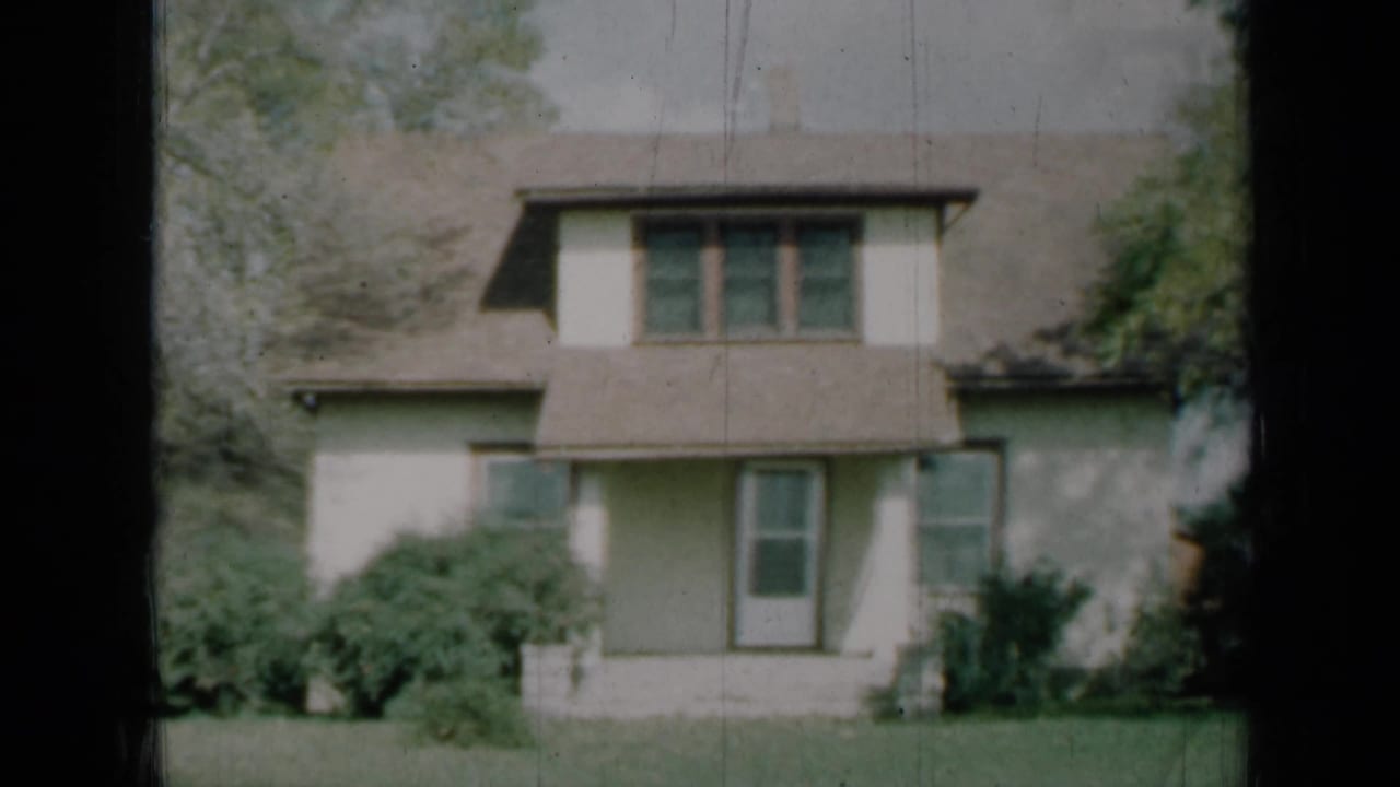 Two-story cream house with brown shingled roof, porch, shrubs, trees, and lawn.