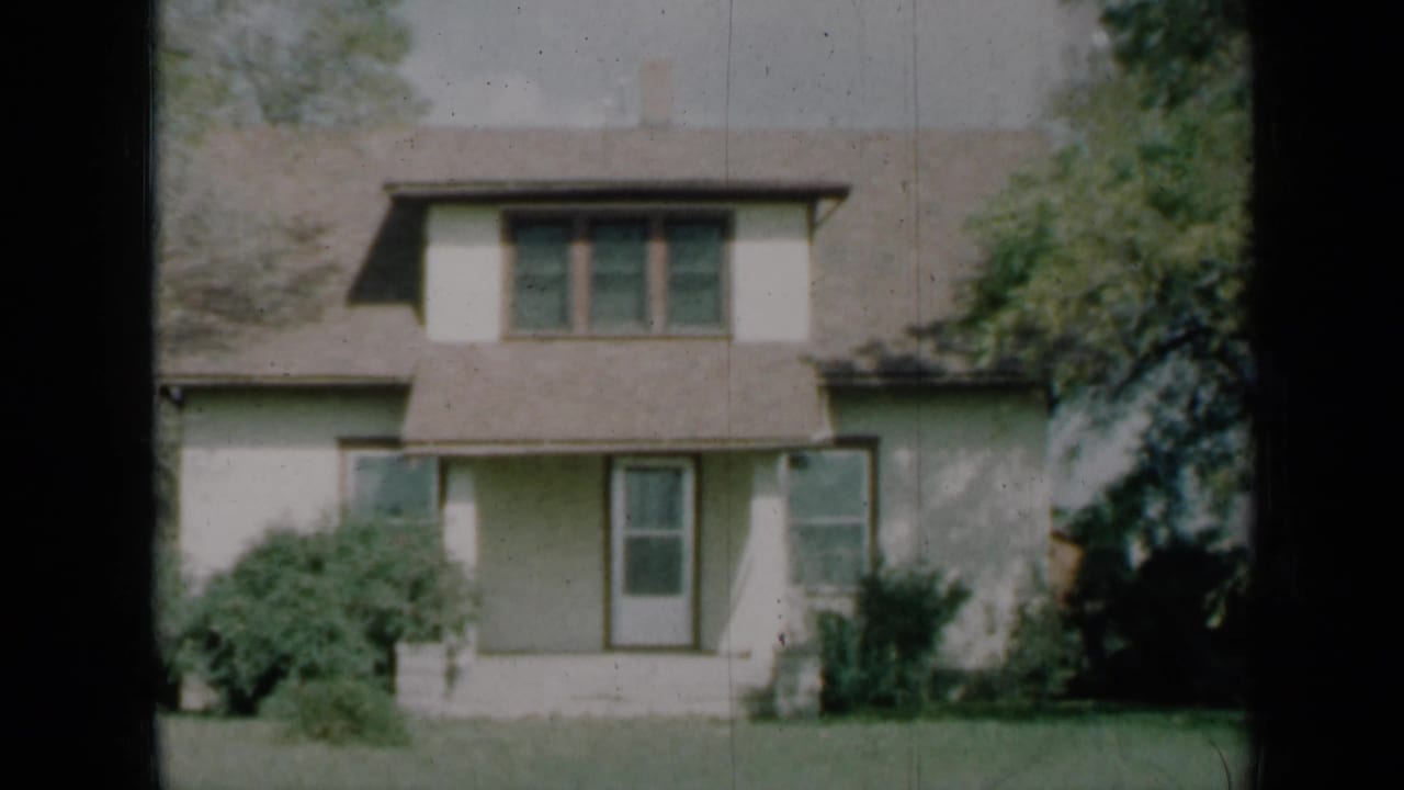 1961 Missouri home-movie frame of a cream two-story house exterior with porch, upper windows, shrubs, trees, and a front lawn.