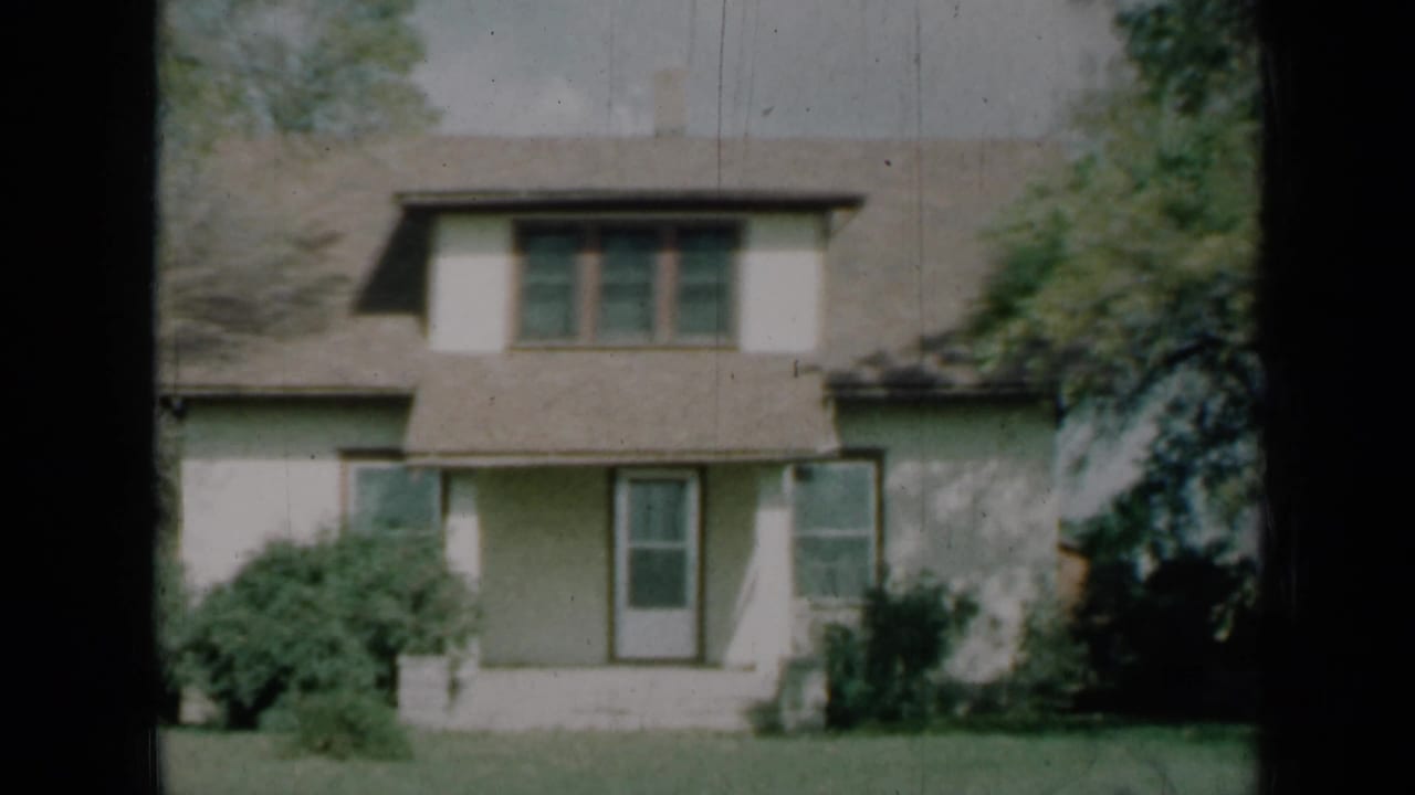 Two-story cream house with dormer windows, central door, shrubs, trees, and lawn.