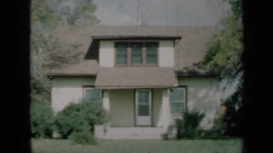 1961 Missouri home-movie still of a cream house facade with upper dormer windows, a centered door, ground-floor windows, shrubs, and lawn.