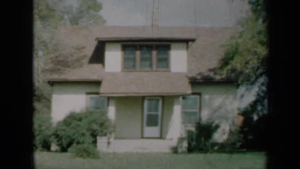 1961 Missouri home-movie frame showing the house front, upper dormer windows, shaded side foliage, shrubs, central door, and lawn.