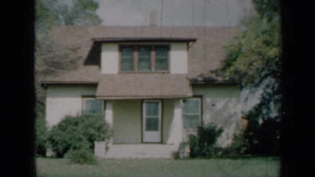 Two-story cream house with brown roof, dormer window, front door, shrubs and lawn.