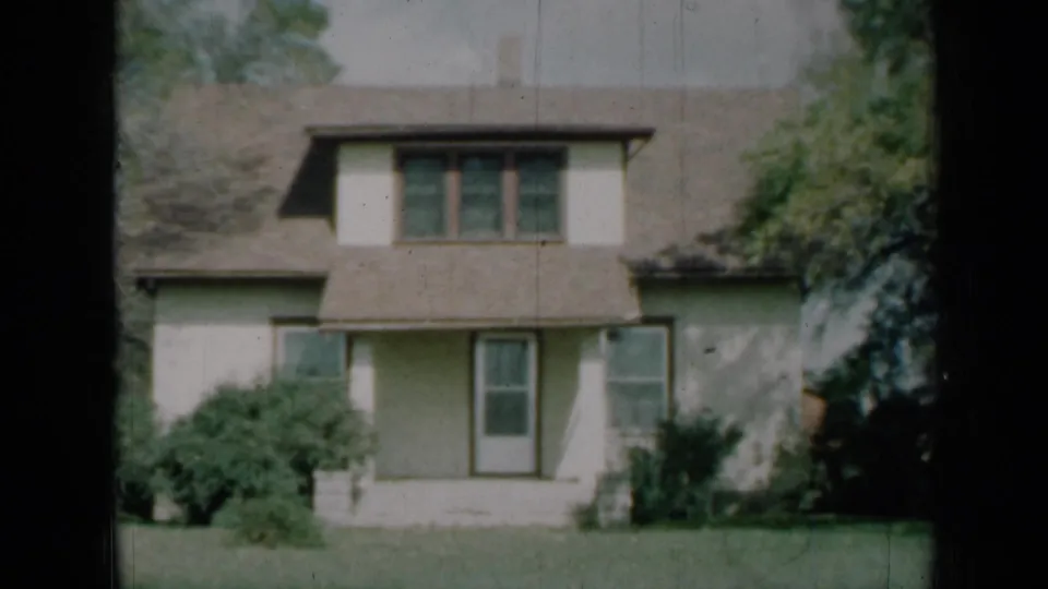1961 Missouri 8mm frame showing a two-story cream suburban house with a brown roof, central dormer, covered entry, shrubs and lawn.