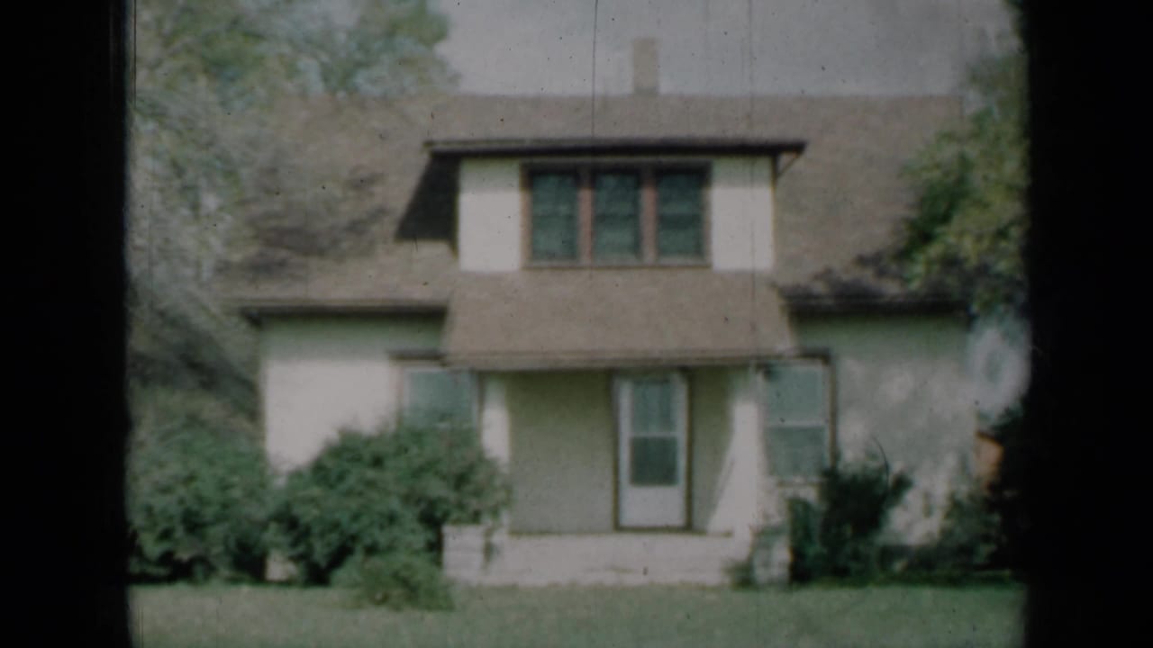 1961 Missouri home-movie frame of the house front, with a central dormer, shaded porch, bushes, lawn and trees at the edges.