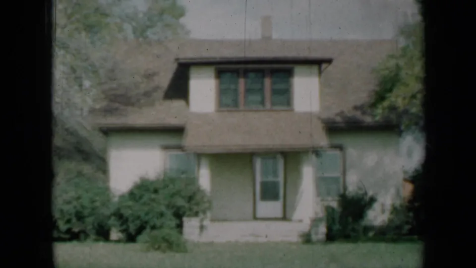 1961 Missouri home-movie frame of the house front, with a central dormer, shaded porch, bushes, lawn and trees at the edges.