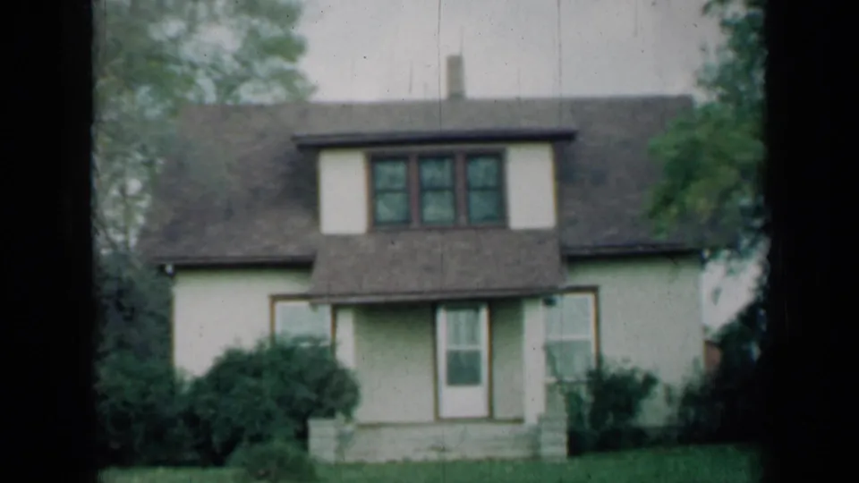 1961 Missouri frame of a cream two-story suburban house with dormer windows, front entrance, bushes, lawn and surrounding tree cover.