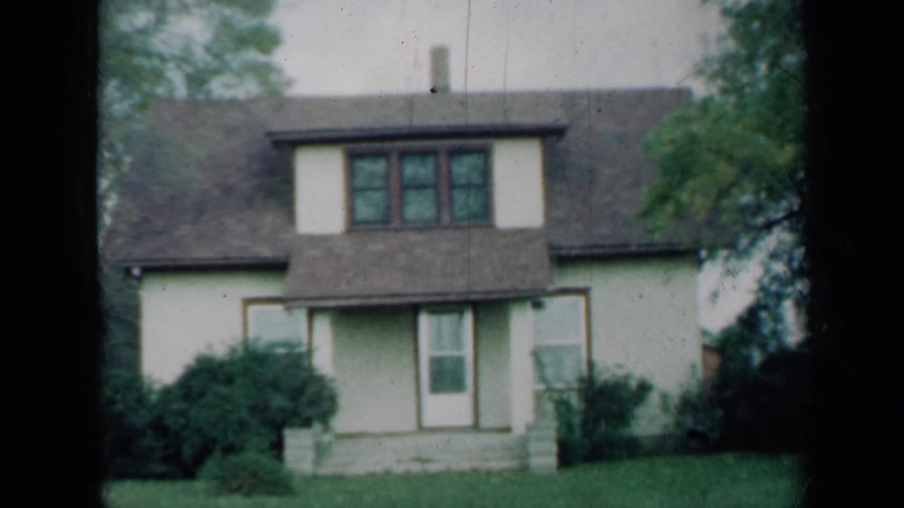 Light-colored two-story house with dormer windows, front door, shrubs, lawn, and tree branches.
