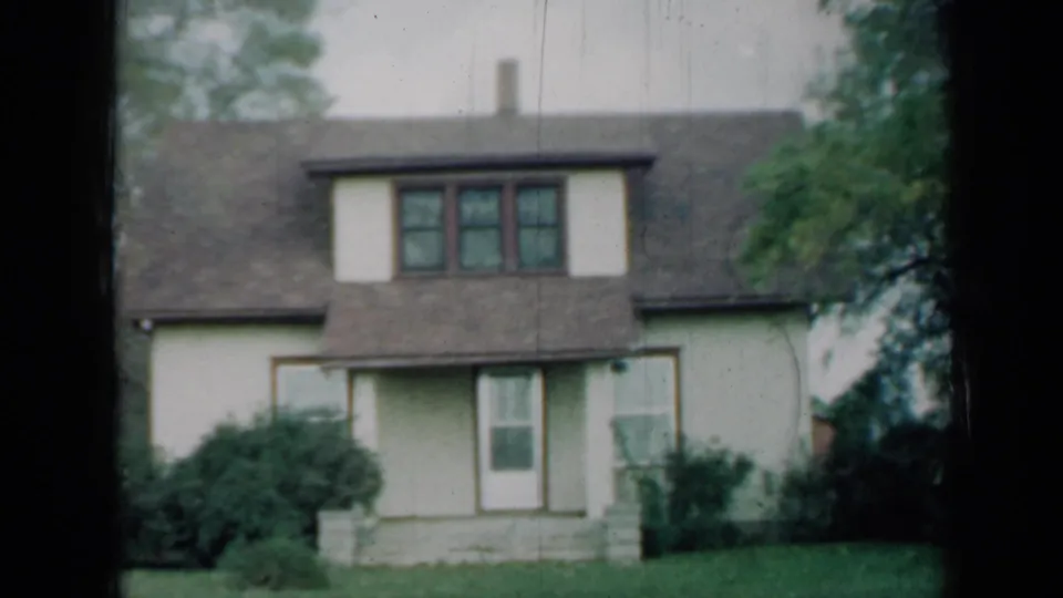 1961 Missouri frame of a quiet residential exterior with dormer windows, a front door, shingled roof, shrubs, lawn, and tree branches.