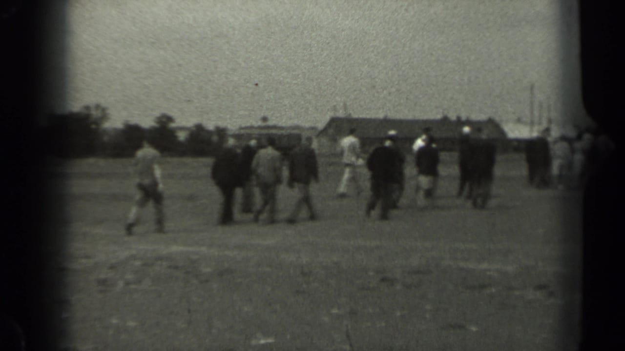 A group of men and other figures walks across an open Missouri field toward low buildings in grainy 1961 home-movie footage.