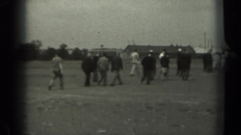 A group of men and other figures walks across an open Missouri field toward low buildings in grainy 1961 home-movie footage.