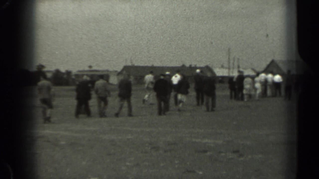 1961 Missouri 8mm frame showing scattered people crossing an open field toward low rural buildings and a barn-like roofline.