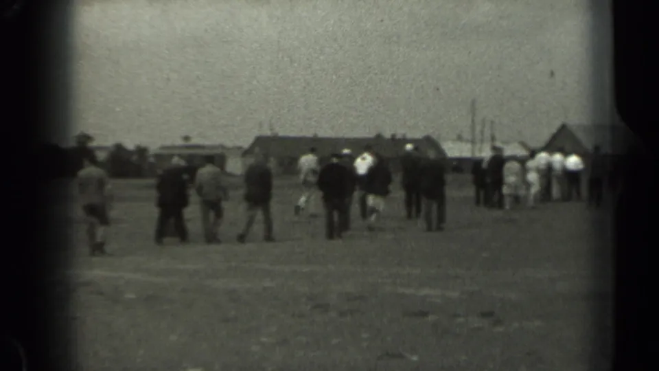 1961 Missouri 8mm frame showing scattered people crossing an open field toward low rural buildings and a barn-like roofline.