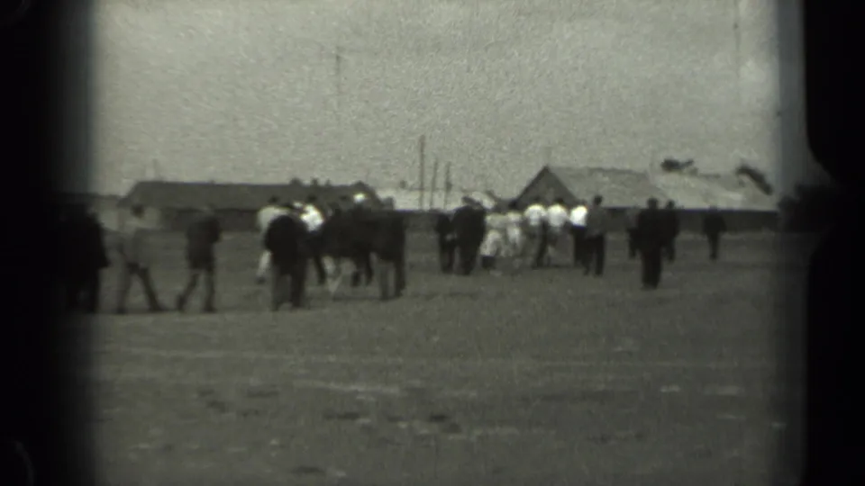 Distant groups in 1961 Missouri appear to walk and gather near low rural buildings across an open field.