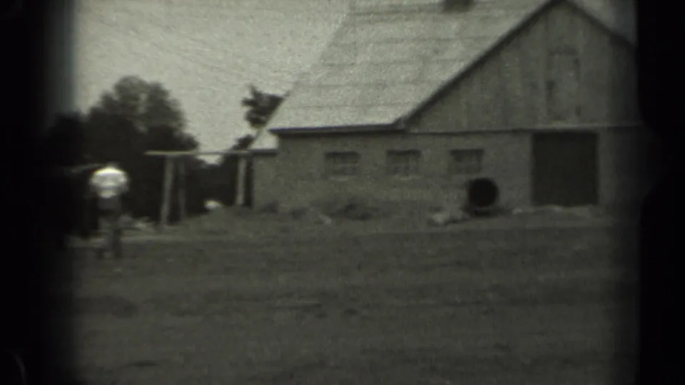 Black-and-white 1961 Missouri farm scene with a barn, open ground, trees, and a distant person near the structures.
